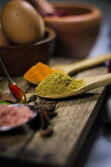 Close up of turmeric powder in wooden spoon surrounded with various asian spices and ingredients on wooden deck.