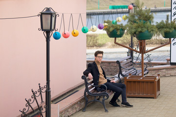 A young man with a pensive face is sitting on a bench. Dressed in a black light overcoat, black-rimmed glasses.