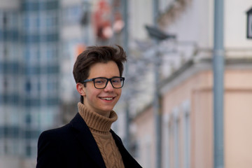 A young man posing with a smile, among the cityscape. Dressed in a black light overcoat, black-rimmed glasses.