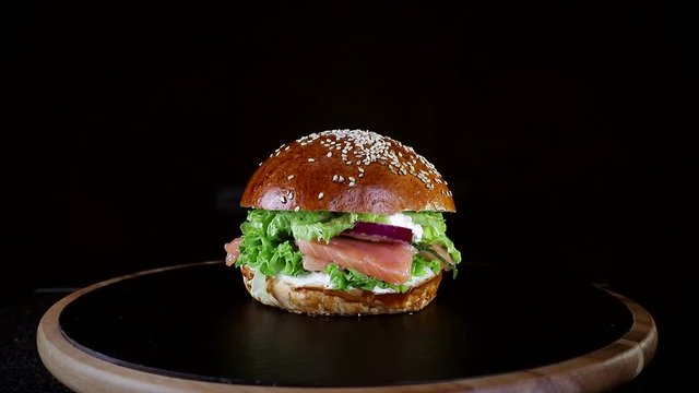Fast food. Burger with red fish, salmon and trout greens, tomato and onion, slowly spinning on a wooden Board on a black background