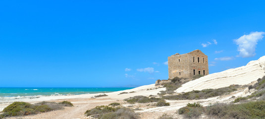 empty white beach with old ruins of abandoned stone house on the rocks and summer blue sky and sea in background in Sicily Italy