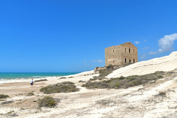 empty white beach with old ruins of abandoned stone house on the rocks and summer blue sky and sea in background in Sicily Italy
