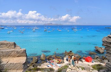 Obraz premium people inside paradise clear torquoise blue water with boats and cloudy blue sky in background in Favignana island, Cala Rossa Beach, Sicily South Italy.