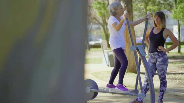 Smiling senior woman training on orbitrek in park. Young female trainer working with elderly client in park. Concept of sport