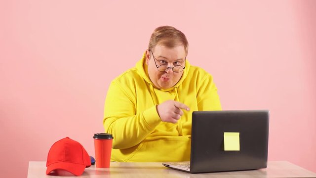 Funny plump man in yellow sweatshirt having foolish face expression using laptop with funny freak grimace