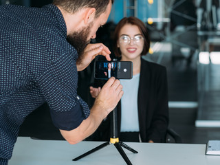 Business workshop. Male assistant interviewing young lady, recording video blog on phone. Vlog...
