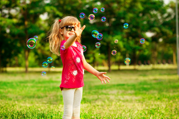 Happy little blond girl blows soap bubbles in summer park