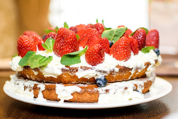 Strawberry cake in cream and mint leaves on the table in a plate, on the background of a teapot with tea, in light colors, close-up with a blurred background.