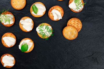 Salty crackers with soft cream cheese spread and herbs, shot from above on a black background with...