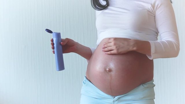 Young unrecognizable pregnant woman tfking care about her stomach skin by applying moisturizing body lotion. Expecting female smoothing her skin with cream.