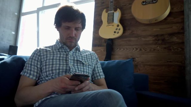 Slowmotion. A man sits on the couch at home and prints a message on the phone connected to the Internet via Wi-Fi. Guiars in the background