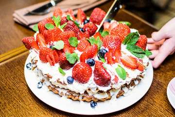 Strawberry cake with blueberries in cream and mint leaves in the process of cutting into pieces on a plate, in light colors, the overall plan