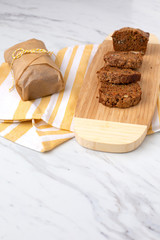 Homemade Zucchini Bread Sliced on Bamboo Cutting Board; Second Loaf Wrapped in Parchment Paper; Yellow and White Dish Towel on White Countertop