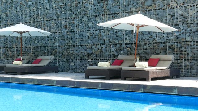 Locked-off shot of sun beds, lounge chairs and parasol on deck next to blue swimming pool, luxury hotel resort.