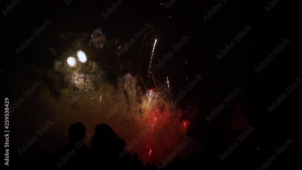 Poster smoke from a fireworks display glows red in the night sky as rockets continue streaking upward and e