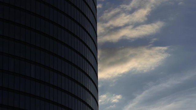 Timelapse with evening clouds flying past steel and glass round-shaped skyscraper corporate office building.