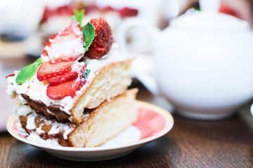A piece of strawberry cake in cream and mint leaves on a table in a plate, in front of a teapot with tea, in light colors, close-up with a blurred background.