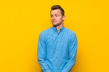 Blonde man over isolated yellow wall making doubts gesture looking side