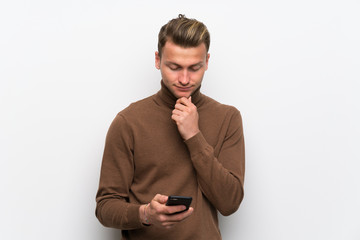 Blonde man over isolated white wall with a mobile and thinking