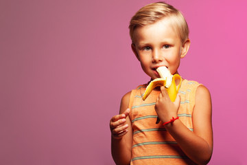 Vegan boy, sweet tooth concept. Funny portrait of smiling boy with blond hair eating yellow banana, posing over pink background. Copy-space. Studio shot