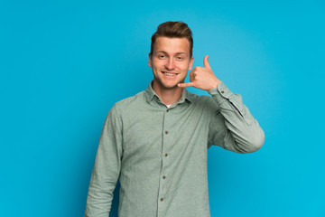 Blonde man over isolated blue wall making phone gesture. Call me back sign