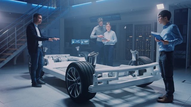 Diverse Automobile Engineers Talking while Working on Electric Car Platform Chassis Prototype. In Automotive Innovation Facility Concept Vehicle Frame Includes Tires, Suspension, Engine and Battery