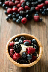 Delicious sweet dessert made from berry mix (raspberries, blueberries and blackberries) with whipped cream in a wooden bowl on a wooden background. Close up.