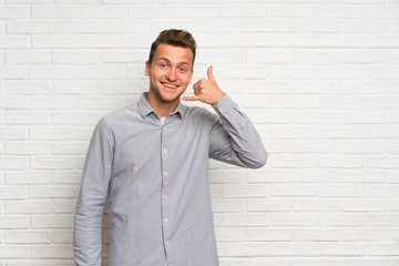 Blonde man over white brick wall making phone gesture. Call me back sign