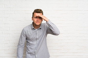 Blonde man over white brick wall looking far away with hand to look something