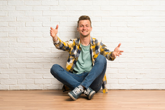 Blonde man sitting on the floor presenting and inviting to come with hand