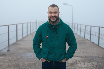 Fall, Winter clothing season concept. Portrait of handsome bearded fisher man wearing marine blue parka, smiling, posing in foggy weather sea scenery. Hands in pockets. Morning time. Outdoor shot