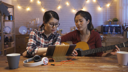 Young happy girl study to play acoustic guitar using tablet computer and having fun while sitting at kitchen table at home in night. two asian women indoor having fun with music using digital pad.