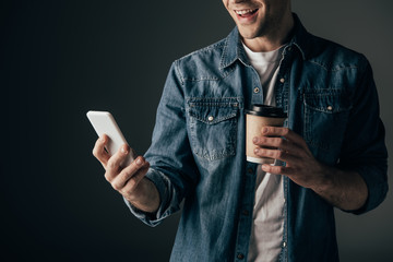 cropped view of man in denim shirt holding paper cup and using smartphone isolated on black