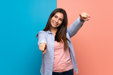 Young woman over pink and blue wall points finger at you while smiling
