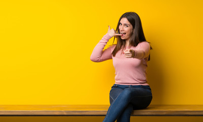Young woman sitting on table making phone gesture and pointing front