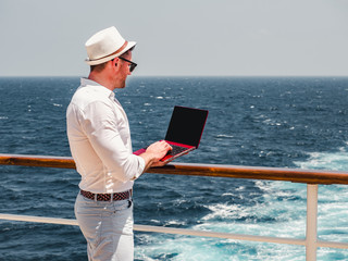 Fashionable man in sunglasses and a sun hat, working on a red laptop on the empty deck of a cruise...