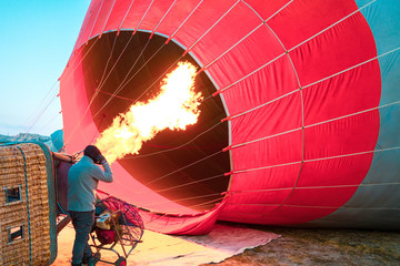 Hot air balloon with flame and basket lying on the ground on the field while filling with air and...