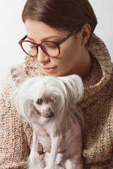 Best girlfriends concept. Young gorgeous lady with natural make-up wearing trendy clothes, eyeglasses kissing small chinese crested dog over white background. Street fashion style. Indoor shot