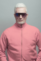 Fabulous at any age, eyewear concept. Portrait of fashionable 60-year-old man in pink jacket over gray background. Trendy haircut, glossy grey hair. Fashionista style. Studio shot