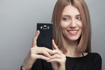 Always online concept. Close up portrait of smiling woman with long chestnut hair, natural make-up, holding smartphone on light gray background. Copy-space. Studio shot