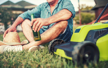 Gardening. Senior man take a rest in the yard and enjoying in beer after work with a lawn mower....
