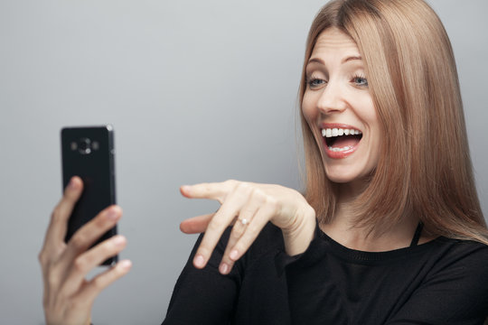 Funny blogger concept. Close up portrait of joking woman with long chestnut hair, natural make-up, holding smartphone, making video call, saying hi on light gray background. Studio shot