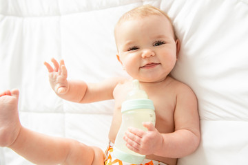 Pretty baby girl drinks milk from bottle lying on bed. Child weared diaper in nursery room.