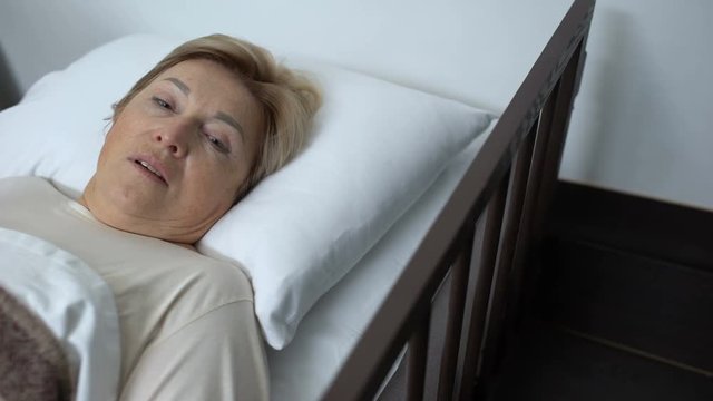Upset senior female lying in hospital bed, touching forehead with trembling hand