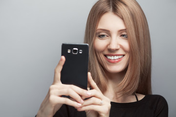Always online concept. Close up portrait of smiling woman with long chestnut hair, natural make-up, holding smartphone on light gray background. Copy-space. Studio shot