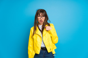 Young woman with yellow jacket on blue background surprised and shocked while looking right