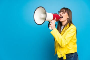 Young woman with yellow jacket on blue background shouting through a megaphone
