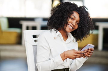 Сheerful business african american lady with afro hair, wear white blouse and green pants posed in cafe at chair and using mobile phone.