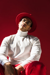 stylish mixed race man in hat and glasses sitting, looking at camera and posing isolated on red