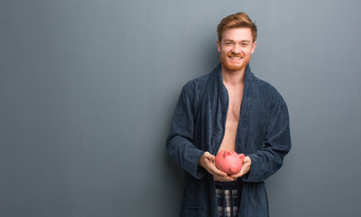Young redhead man wearing pajama cheerful with a big smile. He is holding a piggy bank.